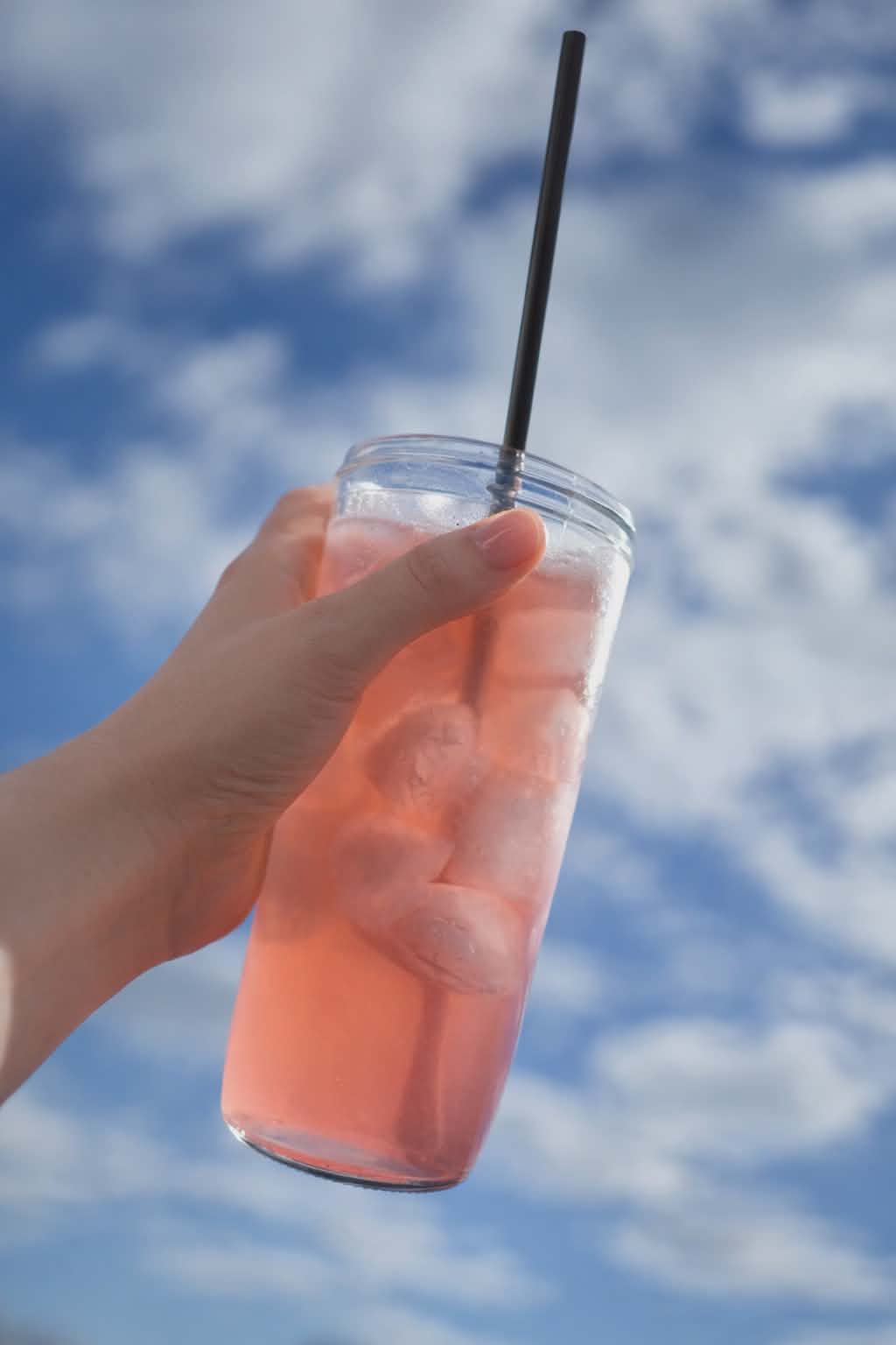 Hand holding a pink iced drink with a straw against a blue sky background