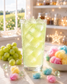 A tall glass of pale lime-green iced loaded tea filled with crystal-clear ice sits on a white wooden countertop in a bright cottage kitchen. Fresh green grapes and colorful pink, blue, and yellow cotton candy surround the drink, with warm fairy lights, sparkling magical accents, and a sunlit garden view creating a whimsical fantasy potion pantry atmosphere.